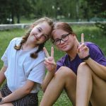 Two cheerful teenage girls sitting on grass, smiling and showing peace signs in a sunny park.