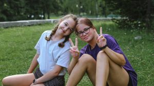 Two cheerful teenage girls sitting on grass, smiling and showing peace signs in a sunny park.