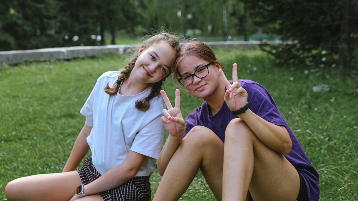 Two cheerful teenage girls sitting on grass, smiling and showing peace signs in a sunny park.